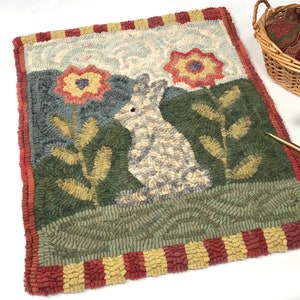 May include: A rectangular rug featuring a hooked fiber design of a white rabbit sitting in a green field with two large yellow and red flowers. The rug has a border of alternating red and yellow blocks and a textured, multi-colored surface.