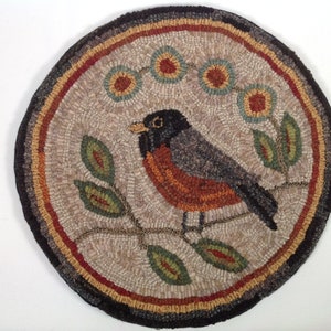 Puede incluir: Alfombra redonda hecha a mano con la imagen detallada de un petirrojo posado en una rama con hojas. La alfombra tiene un fondo neutro y un borde de anillos conc&eacute;ntricos de varios colores. El petirrojo tiene el pecho rojo y las alas grises. Di&aacute;metro aproximado de 60 cm.