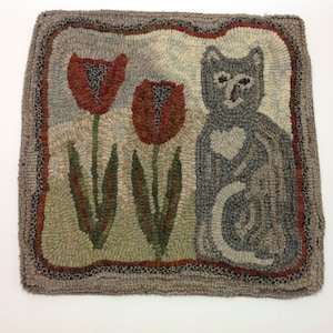 May include: Hand-hooked rug featuring a gray cat with a heart-shaped design and two red tulips. The rug has a neutral color palette with a gray border and green, red, and beige accents. The rug measures approximately 18 inches by 18 inches.