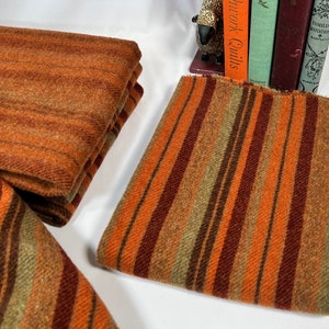 May include: Close-up of folded fabric pieces in shades of orange, brown, and green. One piece has horizontal stripes, while another has vertical stripes. The fabric appears to be a textured wool or similar material.