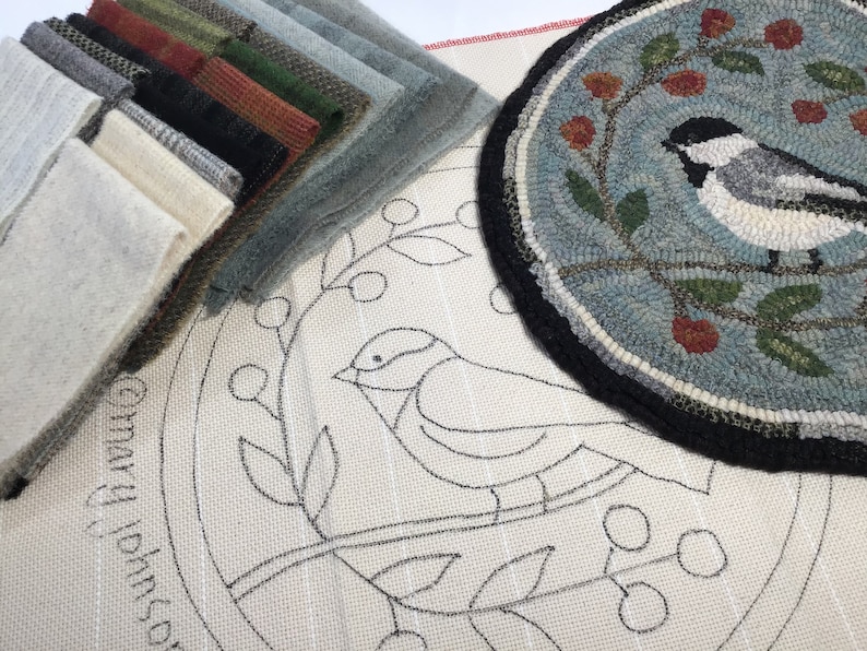 May include: A collection of fabric swatches in various colors, including gray, green, and black, alongside a completed round rug with a bird design. The rug features a black border and a light blue background with floral accents. A canvas with a bird outline is also visible.