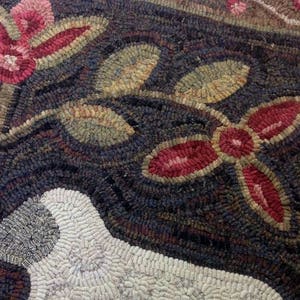 K&ouml;nnte beinhalten: Nahaufnahme eines handgefertigten Teppichs mit einem Schaf, Blumen und Bl&auml;ttern. Der Teppich zeigt ein Schaf in Wei&szlig; mit roten, rosa und gr&uuml;nen floralen Akzenten auf einem dunkelblauen Hintergrund. Der Teppich ist aus Wolle.