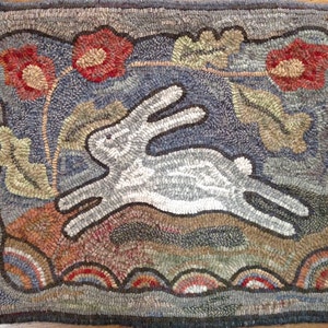 May include: Hand-hooked rug featuring a gray rabbit leaping across a landscape with red poppies and green leaves. The rug has a blue background and a dark gray border. The design includes earth-toned details and a whimsical, folk art style.