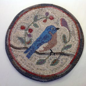 Puede incluir: Una alfombra redonda hecha a mano con un p&aacute;jaro azul posado en una rama con hojas y bayas. El p&aacute;jaro tiene plumas azules y marrones. La alfombra tiene un fondo neutro con un borde azul oscuro y detalles en rojo.