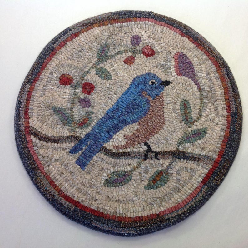 Puede incluir: Una alfombra redonda hecha a mano con un p&aacute;jaro azul posado en una rama con hojas y bayas. El p&aacute;jaro tiene plumas azules y marrones. La alfombra tiene un fondo neutro con un borde azul oscuro y detalles en rojo.