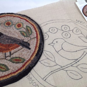 Puede incluir: Una alfombra redonda terminada con un dise&ntilde;o de p&aacute;jaro, con un p&aacute;jaro negro, gris y naranja en una rama con hojas y flores verdes. Junto a ella hay un lienzo con un contorno esbozado del mismo dise&ntilde;o y una peque&ntilde;a tarjeta con el dise&ntilde;o.