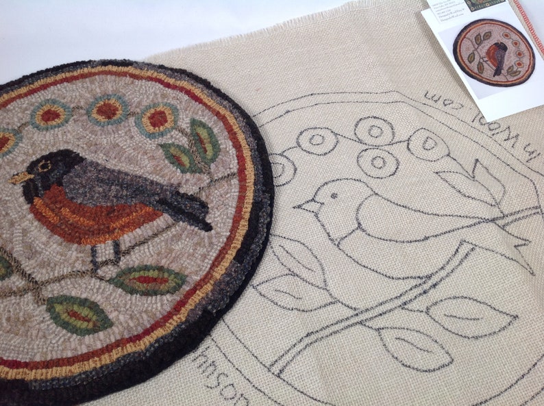 Puede incluir: Una alfombra redonda terminada con un dise&ntilde;o de p&aacute;jaro, con un p&aacute;jaro negro, gris y naranja en una rama con hojas y flores verdes. Junto a ella hay un lienzo con un contorno esbozado del mismo dise&ntilde;o y una peque&ntilde;a tarjeta con el dise&ntilde;o.