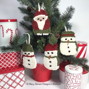 May include: A collection of handmade Christmas ornaments, including Santa Claus and snowman designs, displayed on a small Christmas tree. The ornaments are made of textured wool in red, green, and white. Decorative tins with candy cane and geometric patterns are in the background.