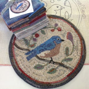 Puede incluir: Una alfombra redonda cosida a mano con un p&aacute;jaro azul posado en una rama con hojas y flores. La alfombra tiene un fondo neutro con un borde oscuro. Una pila de cuadrados de tela de varios colores y una tarjeta con una ilustraci&oacute;n de p&aacute;jaro est&aacute;n en el fondo.