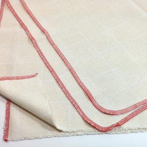 Peut inclure: Tissu blanc cass&eacute; avec un motif quadrill&eacute; et des coutures rouges sur les bords. Le tissu est pli&eacute;, r&eacute;v&eacute;lant la texture et la construction du mat&eacute;riau. Les coutures rouges offrent un d&eacute;tail contrastant.
