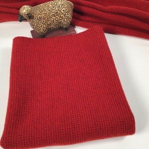 May include: A folded, brick-red knit cowl scarf with a textured pattern. A decorative, gold-colored sheep figurine sits in the background. The scarf is made of a soft, warm material, perfect for cold weather.