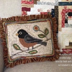 May include: A square, handmade pillow featuring a detailed rug-hooked design of a robin perched on a branch with leaves and a flower. The pillow has a brown ruffled edge and is placed on a neutral-toned sofa. The text "Mary Johnson/Designs in Wool" is visible.