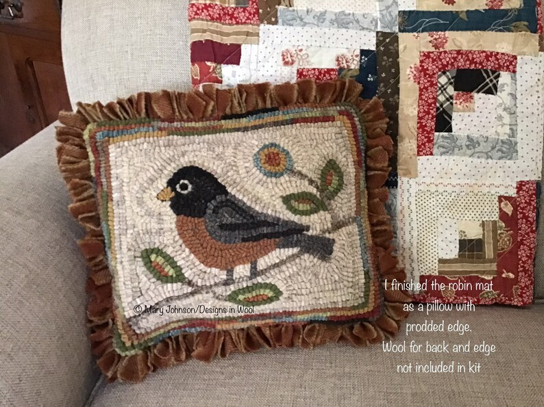 May include: A square, handmade pillow featuring a detailed rug-hooked design of a robin perched on a branch with leaves and a flower. The pillow has a brown ruffled edge and is placed on a neutral-toned sofa. The text "Mary Johnson/Designs in Wool" is visible.