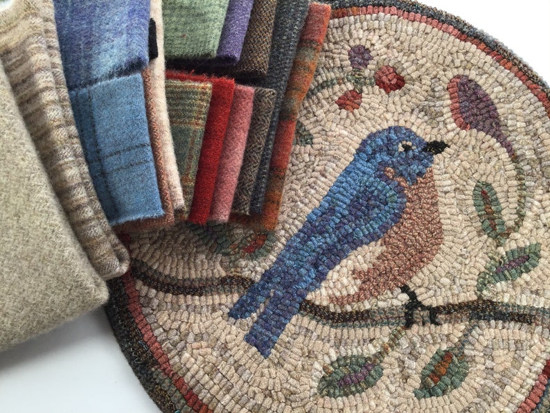 Puede incluir: Una pila de muestras de tela de lana de varios colores junto a una alfombra redonda con un dise&ntilde;o de p&aacute;jaro azul. La alfombra presenta un p&aacute;jaro detallado posado en una rama con hojas y bayas. Las muestras de tela incluyen tonos de azul, verde, rojo y marr&oacute;n.