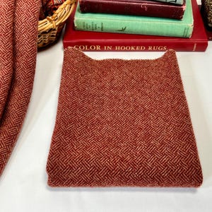 May include: A folded piece of reddish-brown fabric with a woven herringbone pattern. The fabric is a warm, earthy tone, and the weave creates a textured surface. The fabric is likely a textile for home decor or apparel.