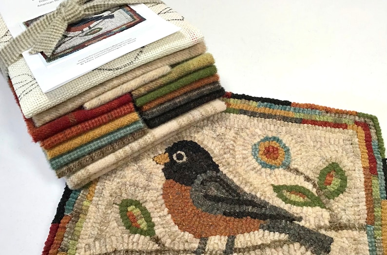 May include: A handcrafted rug with a detailed bird design, featuring a black-headed robin with an orange breast, perched on a branch with a flower. A stack of fabric squares in various colors is tied with a ribbon, complementing the rug's color palette.