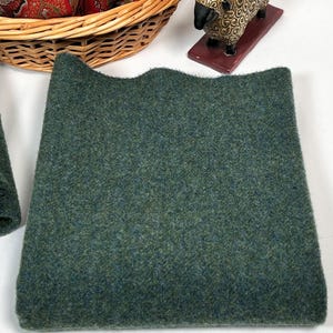 May include: A piece of dark green wool fabric with a scalloped edge. The fabric has a textured appearance and a mix of green and blue fibers. A woven basket and a decorative sheep figurine are in the background.