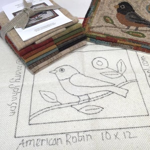 May include: A stack of wool fabric squares in various colors, tied with a beige ribbon, next to a finished rug with a bird design. The rug features a brown bird on a branch with blue and yellow flowers. A larger piece of fabric has a sketched bird design with the text "American Robin 10 x 12".