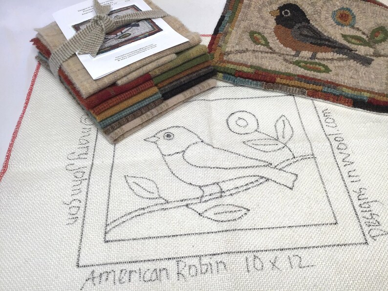 May include: A stack of wool fabric squares in various colors, tied with a beige ribbon, next to a finished rug with a bird design. The rug features a brown bird on a branch with blue and yellow flowers. A larger piece of fabric has a sketched bird design with the text "American Robin 10 x 12".