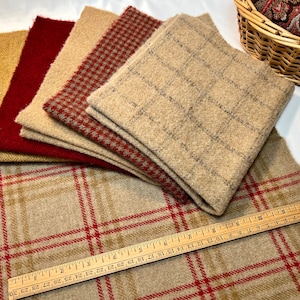 May include: Assortment of fabric swatches in various patterns and colors, including plaid, houndstooth, and herringbone. Colors include tan, red, and gold. A wooden ruler is placed on the bottom swatch. A basket with fabric is in the background.