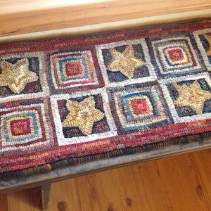 May include: Handmade rectangular rug with a repeating pattern of squares and stars. The squares are multi-colored, with a central square of red, blue, and white. The stars are beige and stitched into the rug. The rug is placed on a wooden bench.