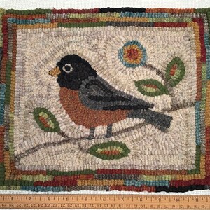 May include: Hand-hooked rug featuring a bird perched on a branch with leaves and a flower. The bird has a black head, gray wings, and a rust-colored chest. The rug has a neutral background with a colorful border. The rug measures approximately 12 inches by 10 inches.