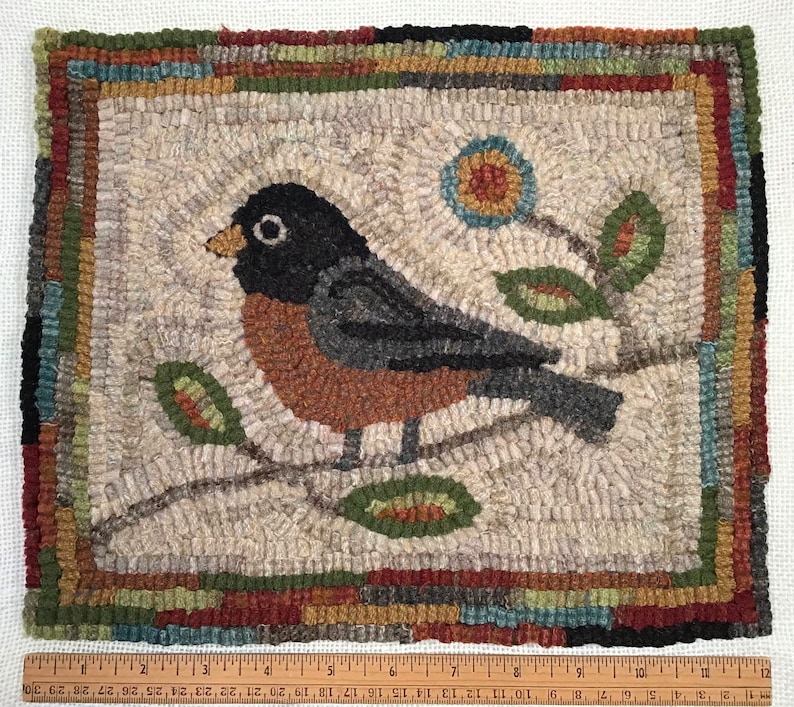 May include: Hand-hooked rug featuring a bird perched on a branch with leaves and a flower. The bird has a black head, gray wings, and a rust-colored chest. The rug has a neutral background with a colorful border. The rug measures approximately 12 inches by 10 inches.