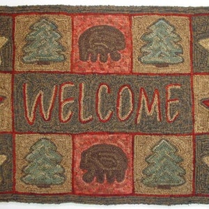 May include: Rectangular welcome mat with a grid pattern. The mat features squares with stars, trees, and bear designs. The word "WELCOME" is stitched in the center. Colors include brown, red, and green.