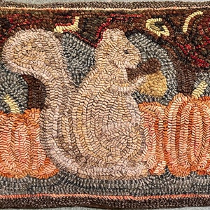 May include: Hand-hooked rug featuring a squirrel holding an acorn between two pumpkins. The squirrel is tan and the pumpkins are orange. The background is a mix of brown, gray, and green.
