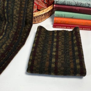 May include: A folded, rectangular textile with a plaid pattern in shades of green, brown, and burgundy. The fabric has a textured appearance. A matching, rolled textile is visible to the left. A stack of vintage books is in the background.