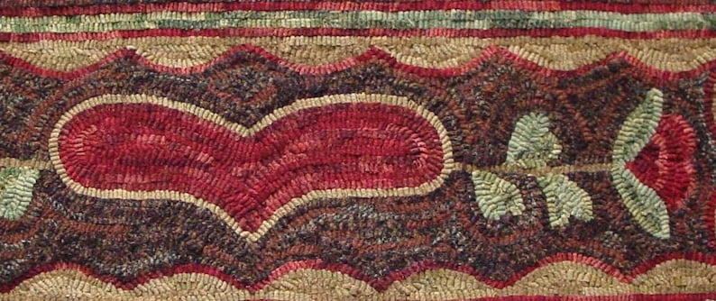 May include: Close-up of a handmade rug with a heart and rose design. The heart is red with a tan border, and the rose is red with green leaves. The background is a mix of brown, tan, and green.