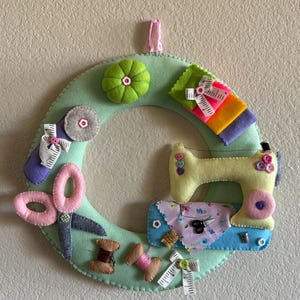May include: A handmade felt sewing wreath in shades of green, pink, yellow, and blue. The wreath features felt representations of sewing tools, including scissors, a sewing machine, spools of thread, and a measuring tape. A pink ribbon is attached for hanging.