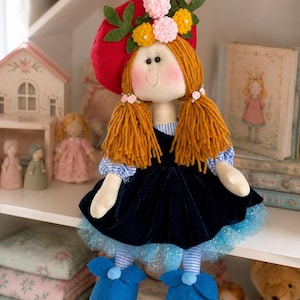Puede incluir: Una muñeca de tela con cabello de lana en coletas, que lleva una boina roja adornada con flores de fieltro, una camisa de rayas azules y blancas, un vestido de terciopelo azul oscuro, una falda de tul azul brillante y zapatos de fieltro azul con lazos pequeños.