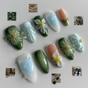 Puede incluir: Una colección de uñas artificiales con varios diseños. Algunas uñas presentan adornos florales en 3D con detalles dorados, mientras que otras tienen colores degradados, detalles de perlas y un diseño de dos tonos. La paleta de colores incluye tonos verdes, azules y melocotón.