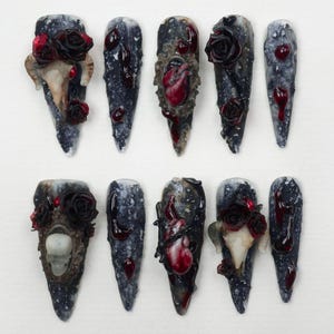 Puede incluir: Un conjunto de diez uñas artificiales largas y puntiagudas con un tema gótico oscuro. Cada uña presenta una base moteada gris y negra, adornada con detalles rojos, rosas y motivos de calaveras o corazones. El diseño general evoca una estética oscura y artística.