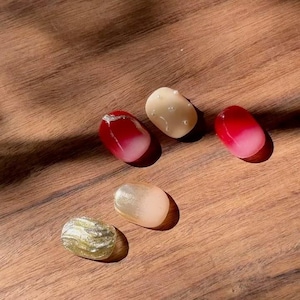 Peut inclure: Cinq pierres ovales polies, dans des tons de rouge, blanc et or, sont disposées sur une surface en bois. Les pierres ont une finition brillante, certaines présentant des accents argentés ou un effet marbré. L'éclairage crée des ombres.