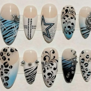 May include: A set of ten artificial nails with various designs. The nails feature patterns like zebra stripes, leopard spots, stars, and swirls, with accents of rhinestones and metallic embellishments. Colors include black, white, blue, and silver.