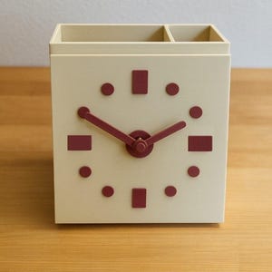 May include: A cream-colored desk organizer with a clock face design. The clock features burgundy-colored hands, dots, and rectangular markers. The top of the organizer has compartments for pens and pencils.