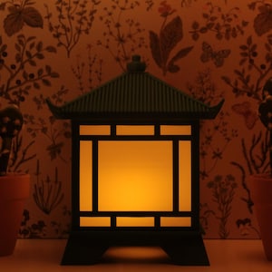 May include: A decorative black lantern with a warm yellow glow, designed in an Asian-inspired style. The lantern is flanked by two terracotta pots with cactus plants. The background features a floral wallpaper.