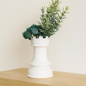 May include: A white ceramic rook chess piece used as a vase, filled with green and teal faux plants. The vase sits on a light wood shelf against a white wall. The plants add a touch of nature.