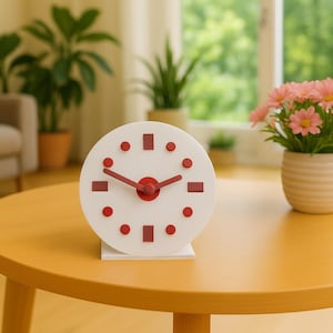 May include: A white, round clock with red accents and hands sits on a yellow table. The clock has red dots and rectangles marking the hours. A small vase of pink flowers is to the right of the clock. The scene is set in a well-lit room.