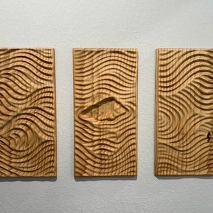 May include: Three rectangular wooden wall art pieces with a carved wave pattern. The wood has a light brown color with visible grain. The middle piece has a carved diamond shape.