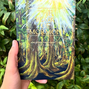 Może przedstawiać: Twarda oprawa książki zatytułowanej "The Book of Mormon" z okładką o tematyce leśnej. Okładka przedstawia żywy obraz lasu z wysokimi, teksturowanymi drzewami i jasnym, żółtym światłem emanującym z góry. Tytuł jest w złotych literach.