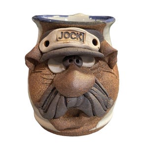 May include: A handmade ceramic mug with a face design. The mug features a brown and tan color scheme, with a sculpted face including a mustache, nose, and a hat that reads "JOCK". The mug has a white interior and a blue rim.