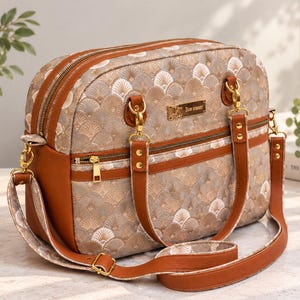 Könnte beinhalten: Eine strukturierte Handtasche mit einem fächerförmigen Muster in Beige und Weiß, braunen Kunstlederbesätzen, goldfarbenen Beschlägen, einem Reißverschluss oben, einer Reißverschlusstasche vorne und einem verstellbaren Schulterriemen.