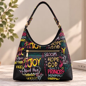 Op de afbeelding: Een zwarte schoudertas in hobo-stijl met een levendige, meerkleurige graffitiprint met woorden zoals JOY, HOPE, GOD, BLOOM en Need Hug. De tas heeft een enkel handvat, een horizontale ritssluiting aan de voorzijde met een goudkleurige trekker.
