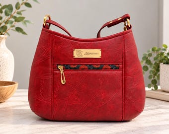 May include: A deep red hobo-style handbag featuring a textured surface with stitched paneling. It includes a gold-toned metal plaque engraved with the text "KS ROWSTREET" and a front zippered pocket with a dark patterned trim and gold hardware.
