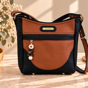 Peut inclure: Un sac bandoulière bicolore noir et marron avec une plaque métallique dorée portant l'inscription Bags by Kasanne. L'avant présente une fermeture éclair avec une breloque composée d'une fleur blanche, d'un chien noir et d'une perle blanche.