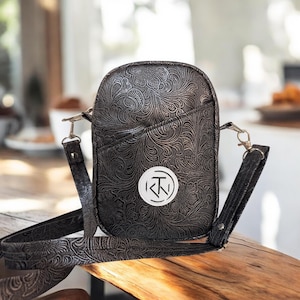 Puede incluir: Bolso bandolera negro con un estampado floral en relieve. El bolso tiene un bolsillo frontal y un parche circular blanco con un logotipo. Incluye una correa ajustable a juego y herrajes plateados.