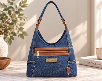 Puede incluir: Un bolso hobo de mezclilla azul con ribetes de cuero marrón, una sola correa para el hombro y un bolsillo frontal con cremallera. El bolso incluye una placa metálica rectangular con el texto "BS BON STREET" y un pequeño detalle de cuero ovalado.